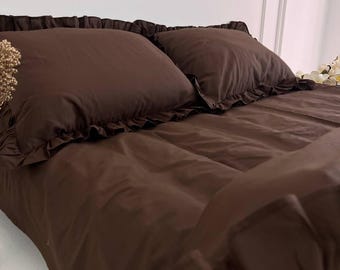 Puede incluir: Un juego de cama de color marrón oscuro sólido que incluye dos fundas de almohada a juego y una funda nórdica. Cada funda de almohada y la funda nórdica tienen un borde decorativo con volantes, lo que aporta una textura uniforme al conjunto.