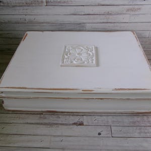 May include: A rectangular white wooden box with a distressed finish showing brown edges. The lid features a centered square relief ornament with a floral motif. The box has a tiered base design and a smooth, flat top surface for display purposes.