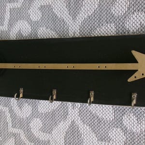 May include: A black wooden key holder with a gold-colored arrow design. The arrow has a detailed head and a long shaft with small holes. Six silver hooks are attached below the arrow.