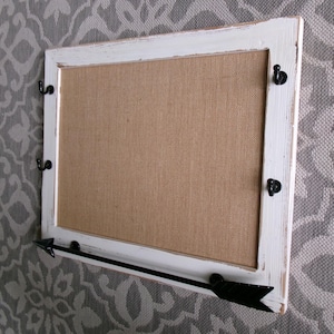 May include: A rectangular wall-mounted display board featuring a distressed white wooden frame and a tan burlap center panel. The frame includes four small black metal hooks on the sides and a black metal arrow-shaped bar along the bottom edge.