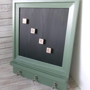 May include: A sage green wooden wall organizer featuring a central black chalkboard surface with four square letter tiles spelling the word LOVE. The unit includes a small horizontal shelf and three silver metal hooks attached to the bottom frame.