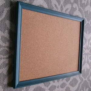 May include: A rectangular cork board featuring a textured, light brown natural cork surface framed by a dark teal-colored border. The frame has a decorative, beaded edge detail running along the inner and outer perimeter of the molding.