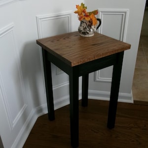 May include: A square side table with a natural wood grain top and four solid black legs. A small ceramic pitcher with a black and white floral pattern sits on the table, containing a decorative arrangement of orange, yellow, and green autumn leaves.