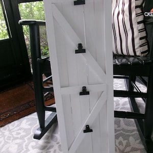 May include: White wooden photo display board with black clips, designed to resemble a barn door. The board has a diagonal brace and is propped up against a black chair with a striped pillow.