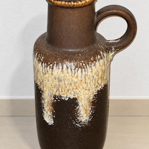 Könnte beinhalten: Eine hohe, zylindrische Keramikvase mit dunkelbrauner Grundfarbe und einem markanten, cremefarbenen Tropfglasur-Effekt im oberen Bereich. Die Vase verfügt über einen einzelnen geschwungenen Henkel am oberen Rand und eine glatte Oberfläche.