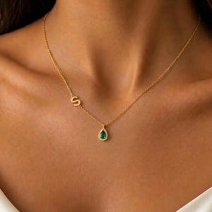 Può includere: Una collana color oro con una catenina sottile, un ciondolo a forma di lettera S posizionato lateralmente e un pendente a goccia con una pietra verde circondata da un alone di piccole pietre trasparenti.