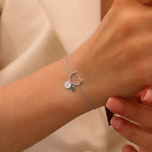 Può includere: Un bracciale a catena color argento caratterizzato da un delicato profilo di testa di gatto, un piccolo ciondolo circolare inciso con la lettera T e una singola pietra preziosa rotonda verde, collegati da una sottile catena.