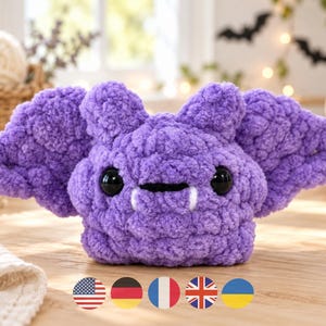 May include: A purple crochet bat plush toy with a textured, fuzzy surface. The bat features two large, round black eyes, a small horizontal mouth, and two white fangs protruding downward. The wings are wide and rounded, extending from the sides.