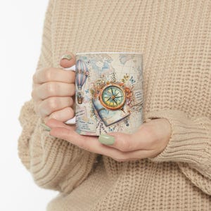 May include: A white ceramic mug featuring a vintage-style travel theme with a world map background, a colorful hot air balloon, a brass-toned compass, and various postage stamp motifs. The design includes text elements like 'PASSENGER TICKET' and 'PARIS'.