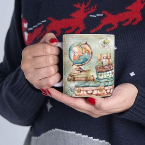 May include: A white ceramic mug featuring a vintage-style illustration of a globe resting on stacked books next to a pile of colorful suitcases. The design includes a small postage stamp graphic and a faded map background with script text.