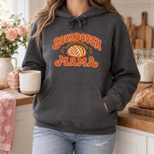 May include: A dark charcoal gray hooded sweatshirt featuring a front kangaroo pocket and a drawstring hood. The chest displays an orange graphic with the text SOURDOUGH MAMA surrounding an illustration of a loaf of scored sourdough bread.