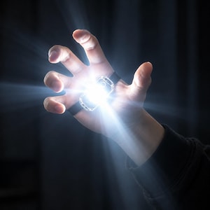 May include: A compact, rectangular LED flashlight secured to the palm with a black elastic strap. The device emits a bright, intense white beam of light that radiates outward, illuminating the surrounding area with a strong, focused glow.
