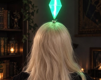 Könnte beinhalten: Ein leuchtender, rautenförmiger Plumbob-Kopfschmuck, der an einer Haarspange befestigt ist. Das durchscheinende, geometrische Objekt strahlt helles Licht aus und sitzt auf langem, welligem blondem Haar, ähnlich einer Videospiel-Figur.