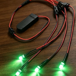 May include: A black battery pack connected to a series of thin red and black electrical wires that terminate in four small, glowing green LED lights. The wires are joined by black plastic connectors, forming a branched circuit assembly.