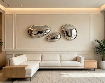 May include: Three irregular, polished silver-toned wall sculptures with a reflective, mirror-like surface are mounted on a beige wall above a cream-colored sectional sofa with integrated light wood side tables and a textured rug.