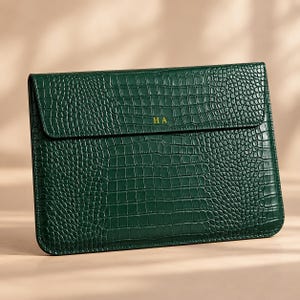 Könnte beinhalten: Eine dunkelgrüne Laptoptasche mit einer strukturierten Krokodilleder-Optik. Das rechteckige Etui verfügt über einen Klappverschluss mit den goldfarbenen, geprägten Buchstaben HA mittig auf der Vorderkante der Klappe.