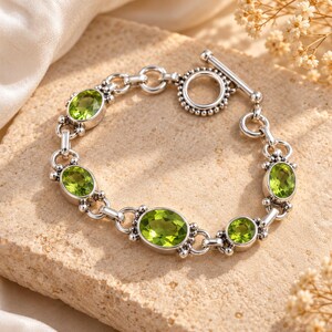 May include: A silver chain bracelet featuring five oval-cut, light green peridot gemstones set in polished metal bezels. The bracelet is secured with a circular toggle clasp and connected by small, round silver links with beaded detailing.