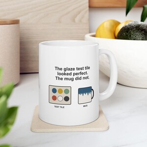 May include: A white ceramic mug featuring the black text 'The glaze test tile looked perfect. The mug did not.' Below the text are two illustrations: a rectangular test tile with six colored circles and a mug with a blue drip glaze pattern.