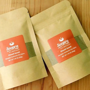 May include: Two brown paper pouches with orange labels. The labels have white text that reads "Soopra It's delicious! Khmeli-Suneli Georgian Authentic Dry Spice NET WT 1.76 OZ (50g)"