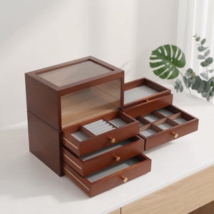 May include: A dark brown wooden jewelry box featuring a glass top lid and multiple pull-out drawers. The interior compartments are lined with light gray fabric, including dedicated ring rolls and square dividers for organizing various small accessories.