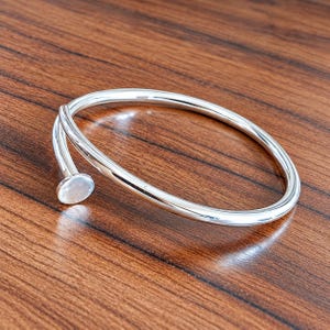 May include: A polished silver-tone metal bangle bracelet featuring a smooth, circular band that curves into an open design. One end of the bracelet terminates in a flat, circular nail-head detail, while the other end tapers to a rounded point.
