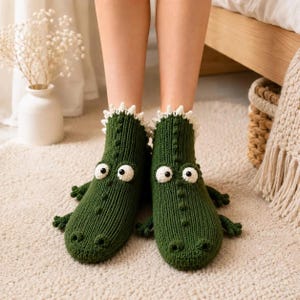 May include: A pair of dark green knit ankle socks designed to resemble crocodiles. The socks feature white crocheted spikes along the back, protruding small limbs on the sides, and large white and black circular eyes on the front.