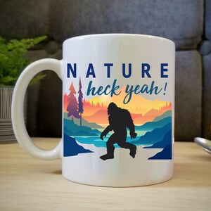May include: White ceramic coffee mug with a colorful nature scene and a silhouette of Bigfoot. The text on the mug reads "NATURE heck yeah!"