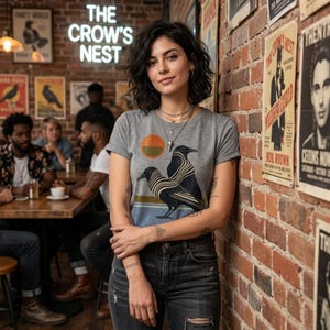 May include: A gray t-shirt with a graphic of two crows and a sun design. The shirt is worn by a person standing in front of a brick wall with posters. The neon sign above reads "THE CROW'S NEST".
