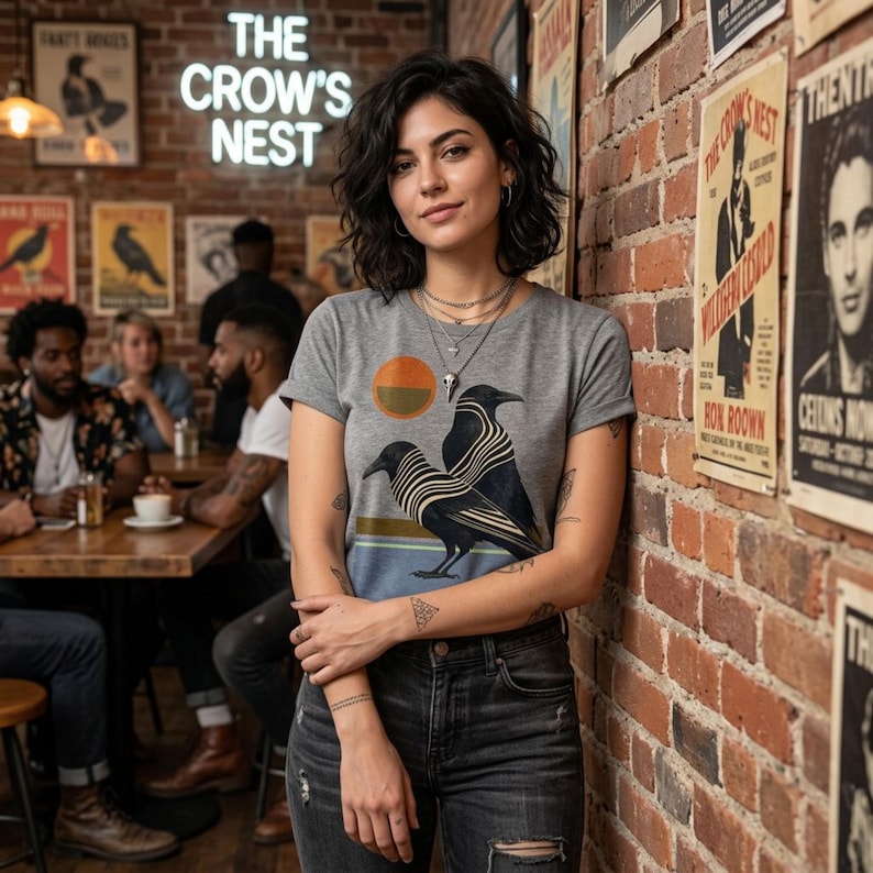 May include: A gray t-shirt with a graphic of two crows and a sun design. The shirt is worn by a person standing in front of a brick wall with posters. The neon sign above reads "THE CROW'S NEST".