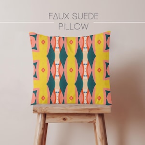 May include: A square faux suede pillow featuring a repeating geometric pattern in vibrant yellow, teal, coral, and cream. The text "FAUX SUEDE PILLOW" is printed in black above the pillow. The design consists of vertical diamond and oval shapes.