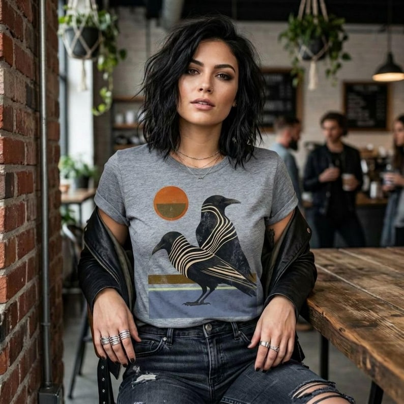 May include: A gray t-shirt featuring a graphic of two black ravens with striped details, a setting sun, and horizontal stripes. The shirt is worn with a black leather jacket and ripped jeans.