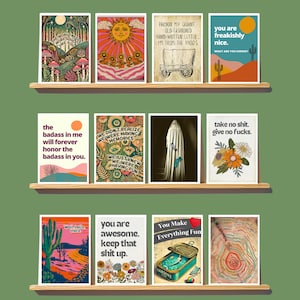 May include: A collection of framed art prints displayed on wooden shelves. The prints feature various designs, including illustrations with text like "The world needs more soul like you" and "You are freakishly nice."
