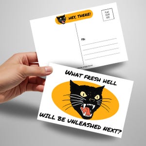 May include: Two white postcards are held in a hand. One postcard has the text "HEY, THERE!" and a black cat illustration. The other postcard has the text "WHAT FRESH HELL WILL BE UNLEASHED NEXT?" and a black cat illustration.