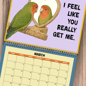 May include: A wall calendar for March featuring a green lovebird perched on a wooden stand looking at its reflection in a heart-shaped mirror. The text reads I FEEL LIKE YOU REALLY GET ME. The calendar grid shows days of the month.