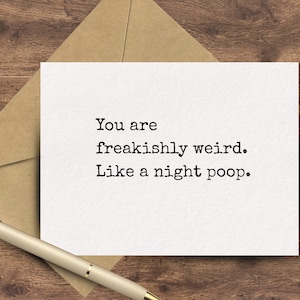 May include: A white greeting card with the text "You are freakishly weird. Like a night poop." is displayed on a wooden surface, next to a gold pen and a brown envelope.