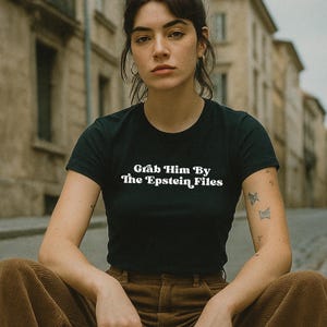 May include: Black crew neck t-shirt with the text "Grab Him By The Epstein Files" in white. The model is wearing brown corduroy pants. The background is a blurred street scene.