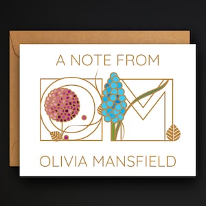 May include: A white card with gold lettering that reads "A Note From Olivia Mansfield". The card features a stylized floral design with a pink flower and a blue flower.