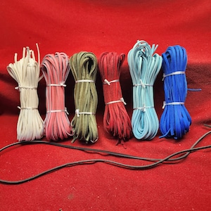 May include: Six spools of leather cord in various colors, including white, pink, green, red, blue, and dark blue, arranged in a row on a red surface. A black leather cord is also visible in the foreground.