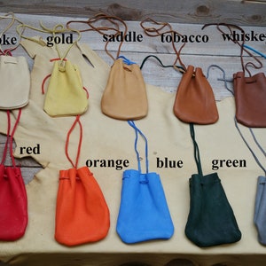 May include: Ten leather pouches in various colors, including smoke, gold, saddle, tobacco, whiskey, red, orange, blue, green, and gray. Each pouch has a drawstring closure and is displayed on a light brown surface.
