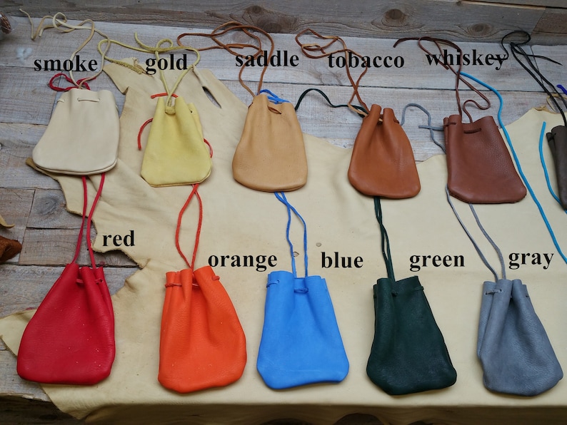 May include: Ten leather pouches in various colors, including smoke, gold, saddle, tobacco, whiskey, red, orange, blue, green, and gray. Each pouch has a drawstring closure and is displayed on a light brown surface.