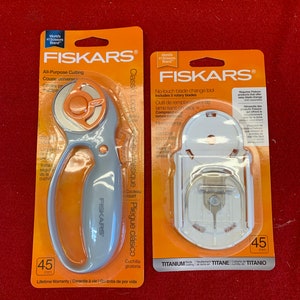 May include: Two Fiskars rotary cutters, one with a classic loop design and the other with a no-touch blade change tool. Both cutters are orange and gray and feature a 45mm blade. The classic loop cutter has a lifetime warranty.