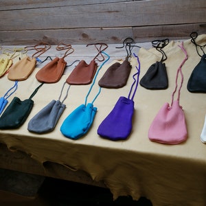 May include: A collection of 15 small leather pouches in various colors, including orange, blue, green, gray, turquoise, purple, pink, white, and brown. The pouches are arranged in a row on a light brown surface.