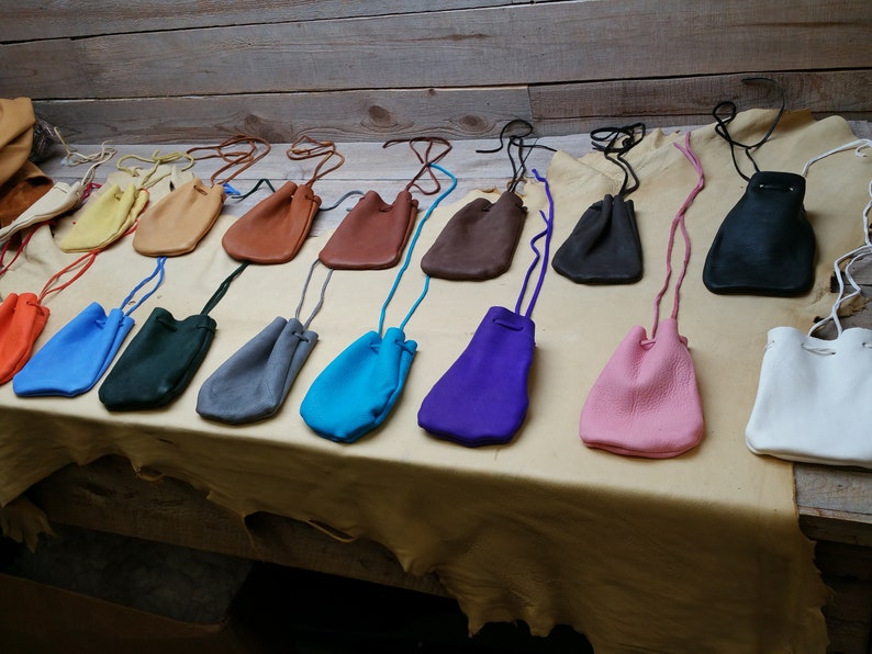 May include: A collection of 15 small leather pouches in various colors, including orange, blue, green, gray, turquoise, purple, pink, white, and brown. The pouches are arranged in a row on a light brown surface.