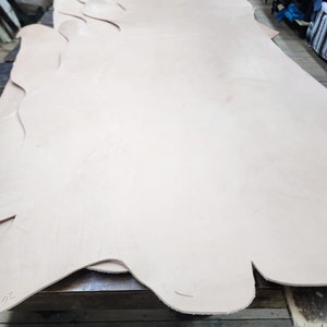 Puede incluir: Una pieza grande de cuero marrón claro, doblada por la mitad y colocada sobre una superficie de madera. El cuero es liso y parece ser de alta calidad.