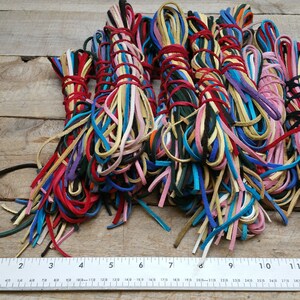 K&ouml;nnte beinhalten: Eine Sammlung bunter Lederkordeln, etwa 30 cm lang, die in B&uuml;ndeln auf einer Holzoberfl&auml;che angeordnet sind. Die Kordeln sind in verschiedenen Farbt&ouml;nen von Rot, Blau, Gr&uuml;n, Gelb, Rosa und Braun.