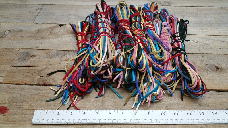 K&ouml;nnte beinhalten: Eine Sammlung bunter Lederkordeln, etwa 30 cm lang, die in B&uuml;ndeln auf einer Holzoberfl&auml;che angeordnet sind. Die Kordeln sind in verschiedenen Farbt&ouml;nen von Rot, Blau, Gr&uuml;n, Gelb, Rosa und Braun.