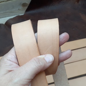 May include: Two strips of light brown leather are held in a hand. The leather is smooth and appears to be of good quality.