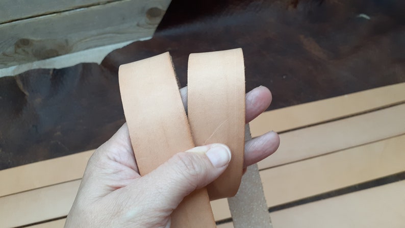 May include: Two strips of light brown leather are held in a hand. The leather is smooth and appears to be of good quality.