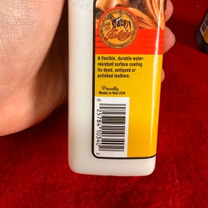 May include: A white bottle with a yellow label that says "Since 1895 Bosine". The label also says "A flexible, durable water-resistant surface coating for dyed, antiqued or polished leathers." and "Proudly Made in the USA". There is a barcode on the label.