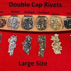 May include: Assortment of double cap rivets in various finishes, including antique copper, nickel plate, antique nickel, antique brass, brass, and gun metal. The rivets are arranged in a wooden tray and displayed on a red surface. The text "Double Cap Rivets" and "Large Size" is visible in the image.
